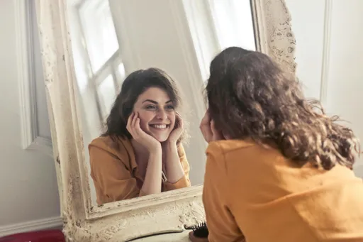 A smiling woman gazing at her own reflection in the mirror while holding her head in her hands
